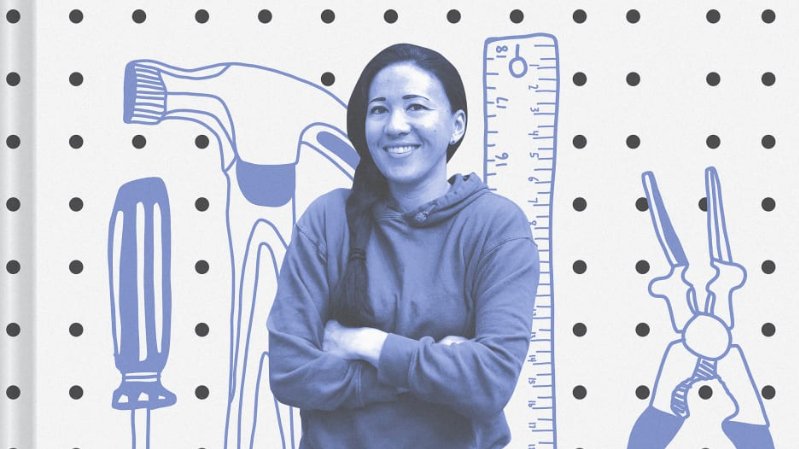 Image: Blue-toned monochrome photo of a woman smiling with braided black hair wearing a hoodie and crossed arms. Background includes part of the front cover of the book, Girls Garage: How to Use Any Tool, Tackle Any Project, and Build the World You Want to See 